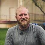A cheerful bearded man smiling outdoors in a casual setting with greenery.