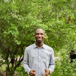 A cheerful man in a shirt smiles in a lush green outdoor setting.